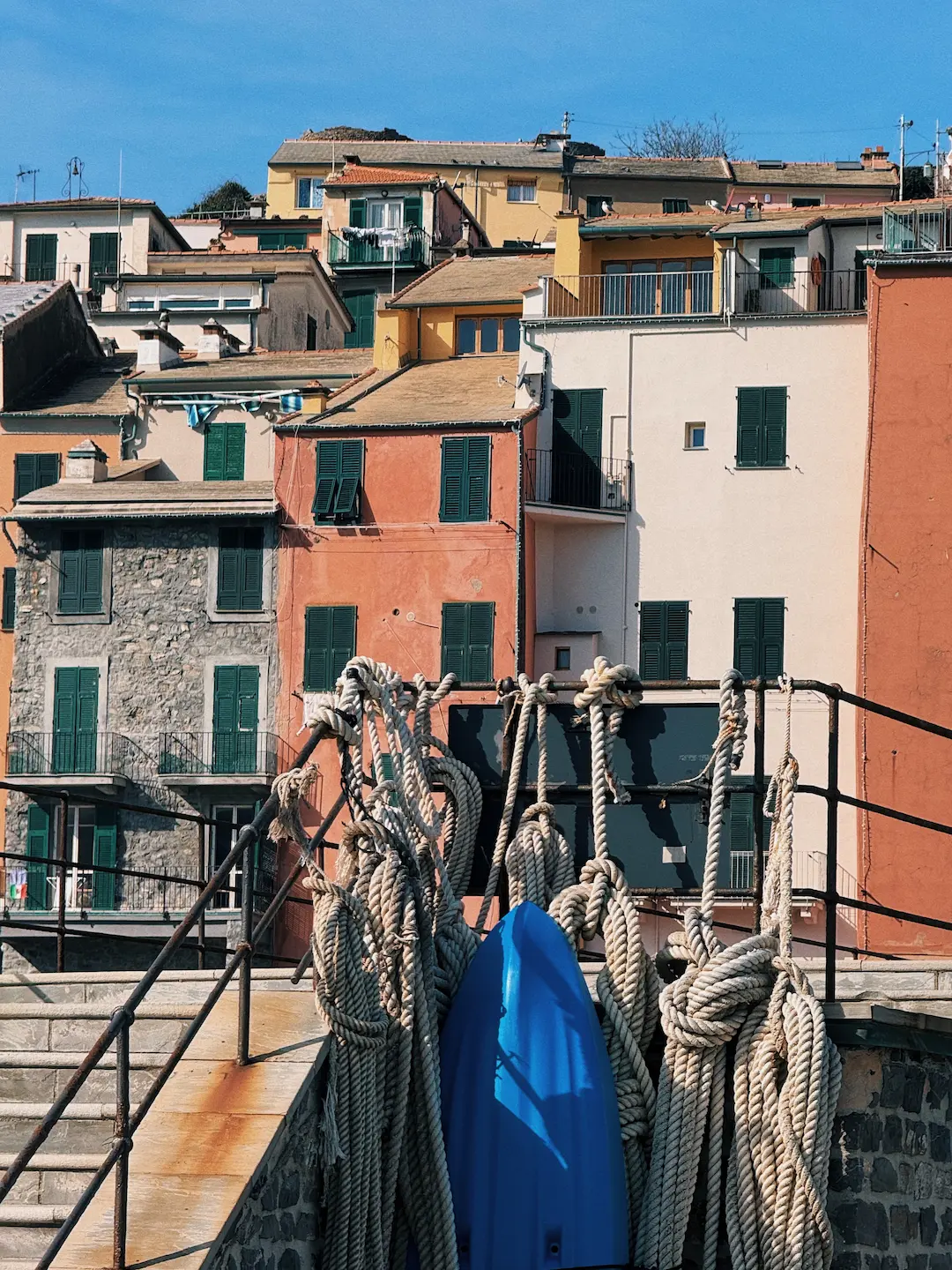 Grosse corde nautiche annodate pendono da una ringhiera metallica arrugginita, con una tavola blu appoggiata dietro. Sullo sfondo, case colorate con persiane verdi si arrampicano lungo la collina sotto un cielo limpido.