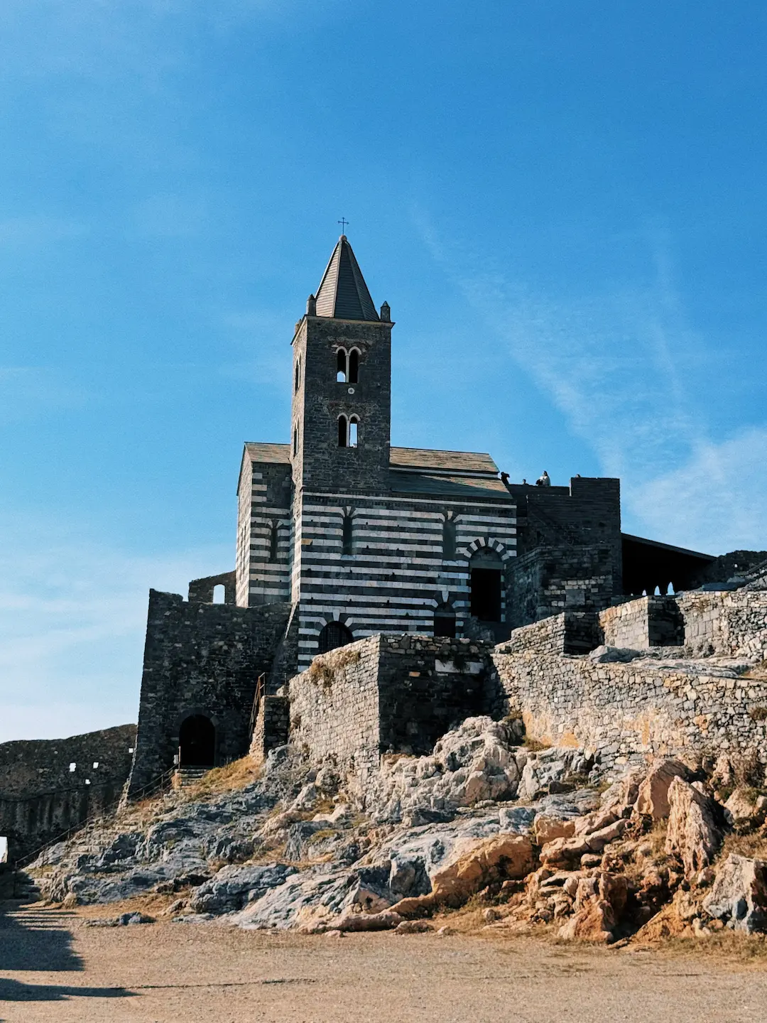 Una chiesa in pietra con facciata a strisce bianche e nere sorge su una scogliera rocciosa. Il campanile svetta contro il cielo azzurro, mentre le antiche mura la circondano, integrandosi con il paesaggio.