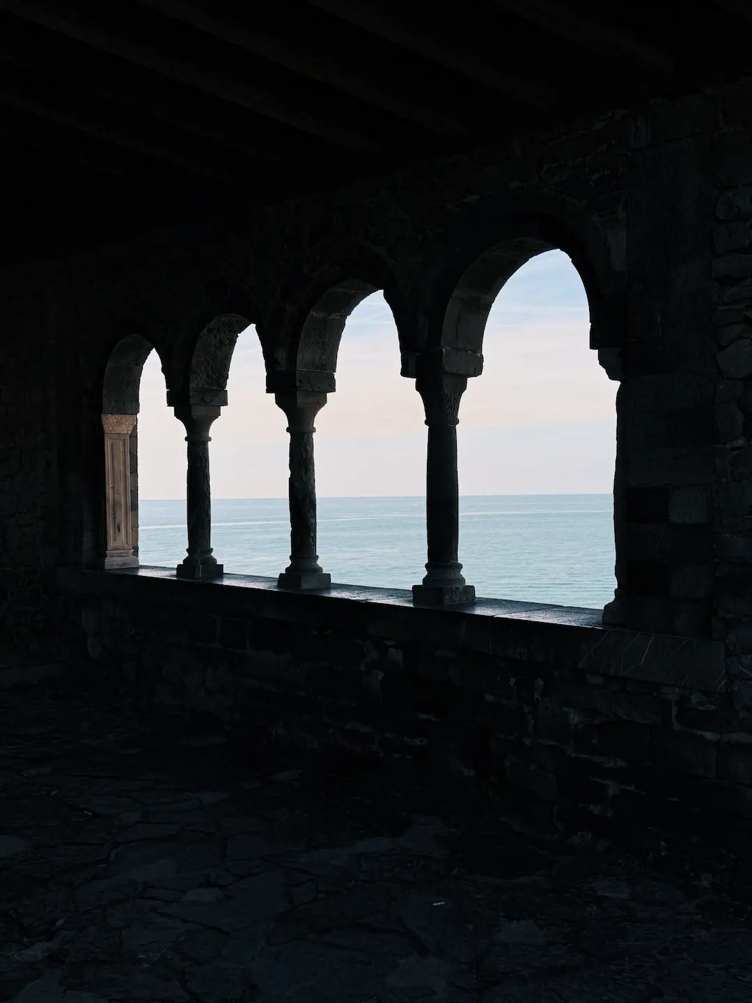 Vista dal buio interno di una struttura in pietra antica verso il mare attraverso quattro archi medievali. Il contrasto tra l'oscurità interna e il panorama luminoso dell'orizzonte marino crea un'atmosfera contemplativa.