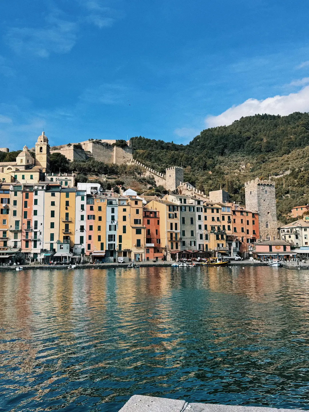 Panorama di Porto Venere visto dal mare, con le caratteristiche case colorate allineate lungo la costa. In primo piano l'acqua riflette i colori degli edifici, mentre sullo sfondo si intravedono la chiesa con campanile, antiche mura e torri medievali che risalgono la collina boscosa sotto un cielo azzurro.