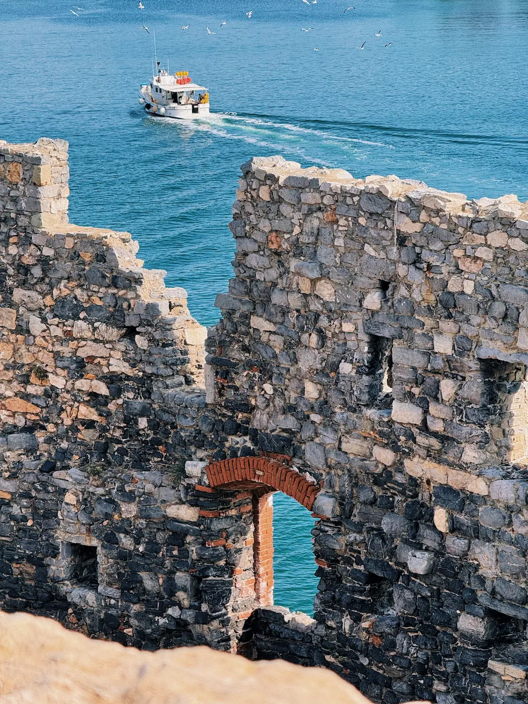 Rovine di un'antica struttura in pietra con un arco in mattoni rossi che incornicia il mare azzurro. In lontananza, un peschereccio bianco naviga sull'acqua seguito da gabbiani. Le mura in pietra grigia mostrano i segni del tempo, creando un suggestivo contrasto tra le antiche vestigia e il paesaggio marino.