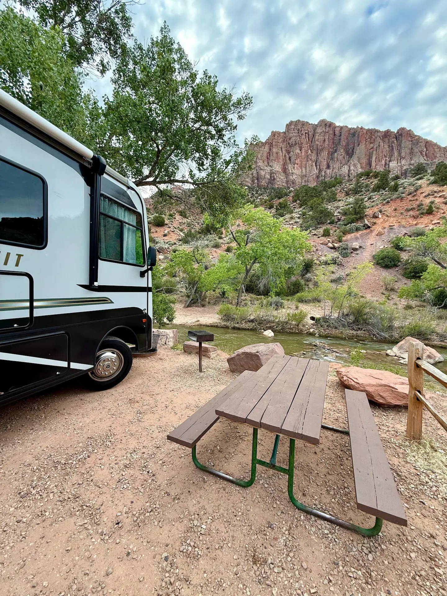 Piazzola del campeggio in un paesaggio naturale, con tavolo da picnic e barbecue vicino a un ruscello, circondati da alberi e rocce rosse tipiche del sud-ovest americano.